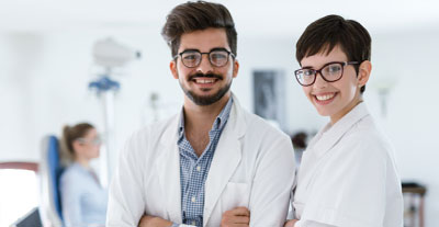 Ein Orthoptist und eine Orthoptistin mit Brille und weißem Kittel lächeln 